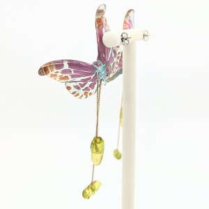 May include: A pair of dangle earrings featuring a butterfly design with pink, white, and orange accents. The butterfly is attached to a gold chain with green glass beads.
