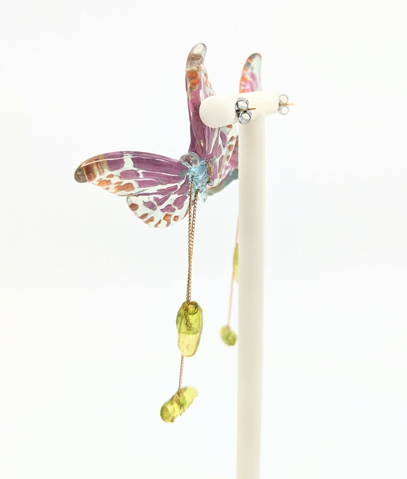 May include: A pair of dangle earrings featuring a butterfly design with pink, white, and orange accents. The butterfly is attached to a gold chain with green glass beads.