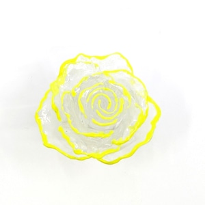 Puede incluir: Una figura de rosa de vidrio transparente con bordes pintados en amarillo brillante en cada pétalo. La flor está formada por pétalos curvos y superpuestos que se enroscan hacia el centro, creando una forma floral dimensional con un contorno amarillo.