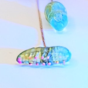 May include: Two blue and green glass beads with pink accents hanging from a gold chain. The beads are shaped like a teardrop and a caterpillar.
