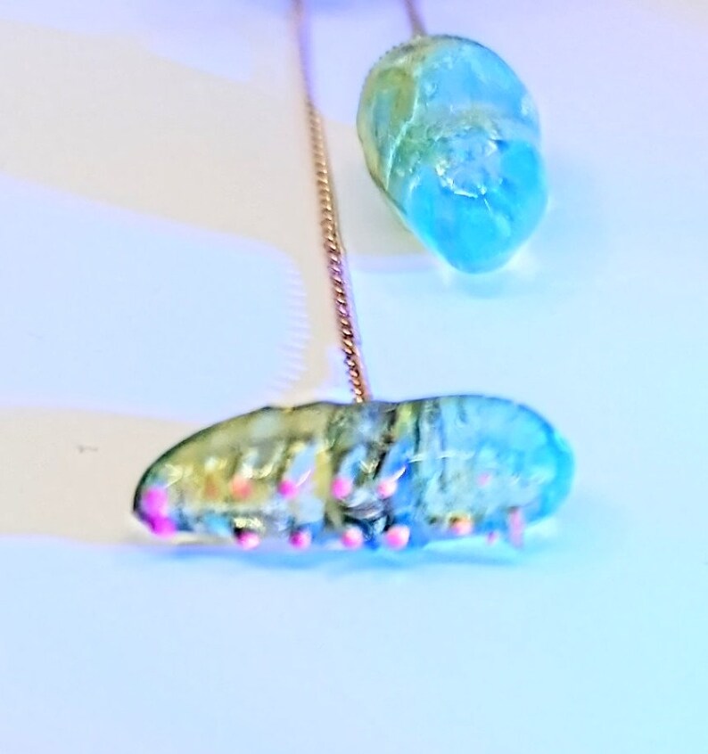 May include: Two blue and green glass beads with pink accents hanging from a gold chain. The beads are shaped like a teardrop and a caterpillar.