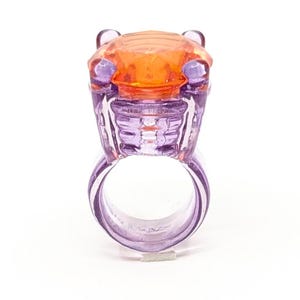 Puede incluir: Un anillo de plástico morado con una parte superior en forma de gema naranja. El anillo está diseñado para parecerse a una cabeza de animal estilizada.