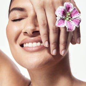 May include: A close-up of a person with a large flower ring. The ring is a white and pink lily with green accents. The person's hand is covering their face, and they are smiling with their eyes closed.