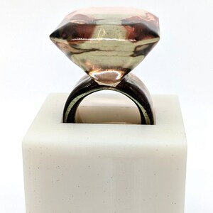 May include: A ring with a large, faceted, clear diamond-shaped stone, set on a brown band. The ring is displayed on a white square base with the text "Marcela Lopez Art" inscribed on the front.
