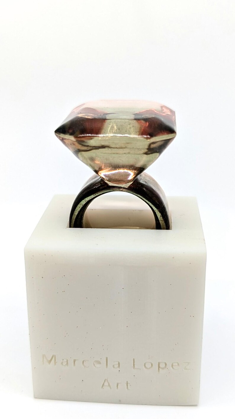 May include: A ring with a large, faceted, clear diamond-shaped stone, set on a brown band. The ring is displayed on a white square base with the text "Marcela Lopez Art" inscribed on the front.