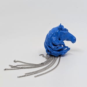 Puede incluir: Una escultura de cabeza de caballo azul con melena fluida y detalles de cadena plateada. La escultura es de un tono azul vibrante y tiene una superficie texturizada. Cadenas plateadas caen de la parte inferior.