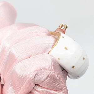 May include: A white ceramic ring with gold polka dots and gold trim. The ring has a small, decorative, gold-colored snail on top. The ring is held by a pink satin fabric.