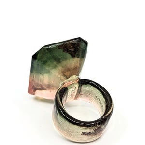 May include: A ring with a large, faceted, green, pink, and black gemstone. The ring is made of ceramic and has a glossy finish.