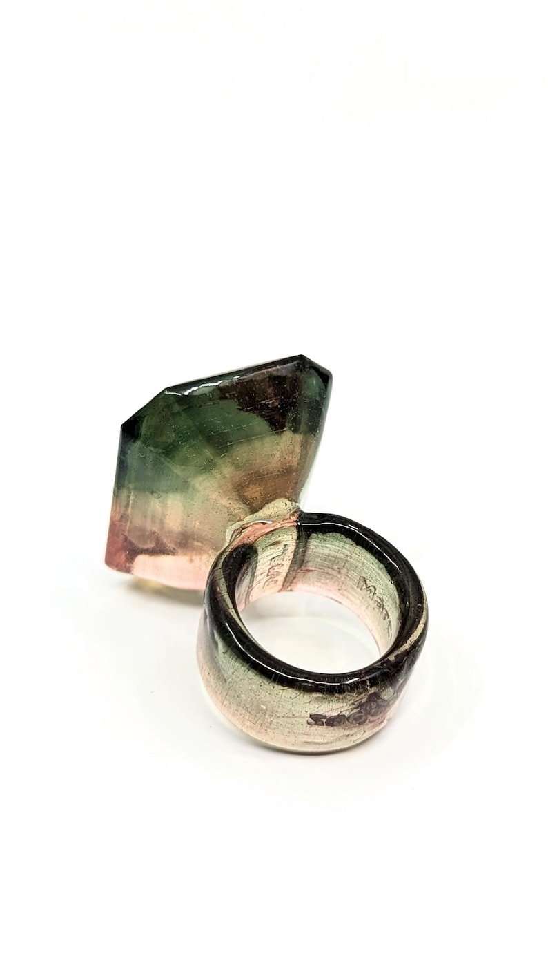 May include: A ring with a large, faceted, green, pink, and black gemstone. The ring is made of ceramic and has a glossy finish.