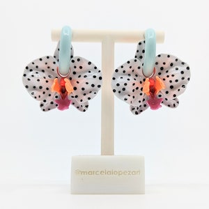 May include: A pair of white orchid earrings with black polka dots and a light blue hoop. The earrings are on a white display stand. @marcelalopezart