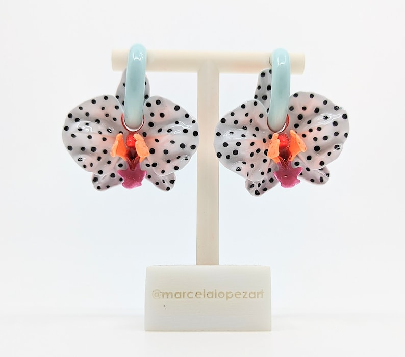 May include: A pair of white orchid earrings with black polka dots and a light blue hoop. The earrings are on a white display stand. @marcelalopezart