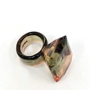 May include: A ring with a large, faceted, clear, green, and pink gemstone. The ring band is a thick, clear, green, and pink band.