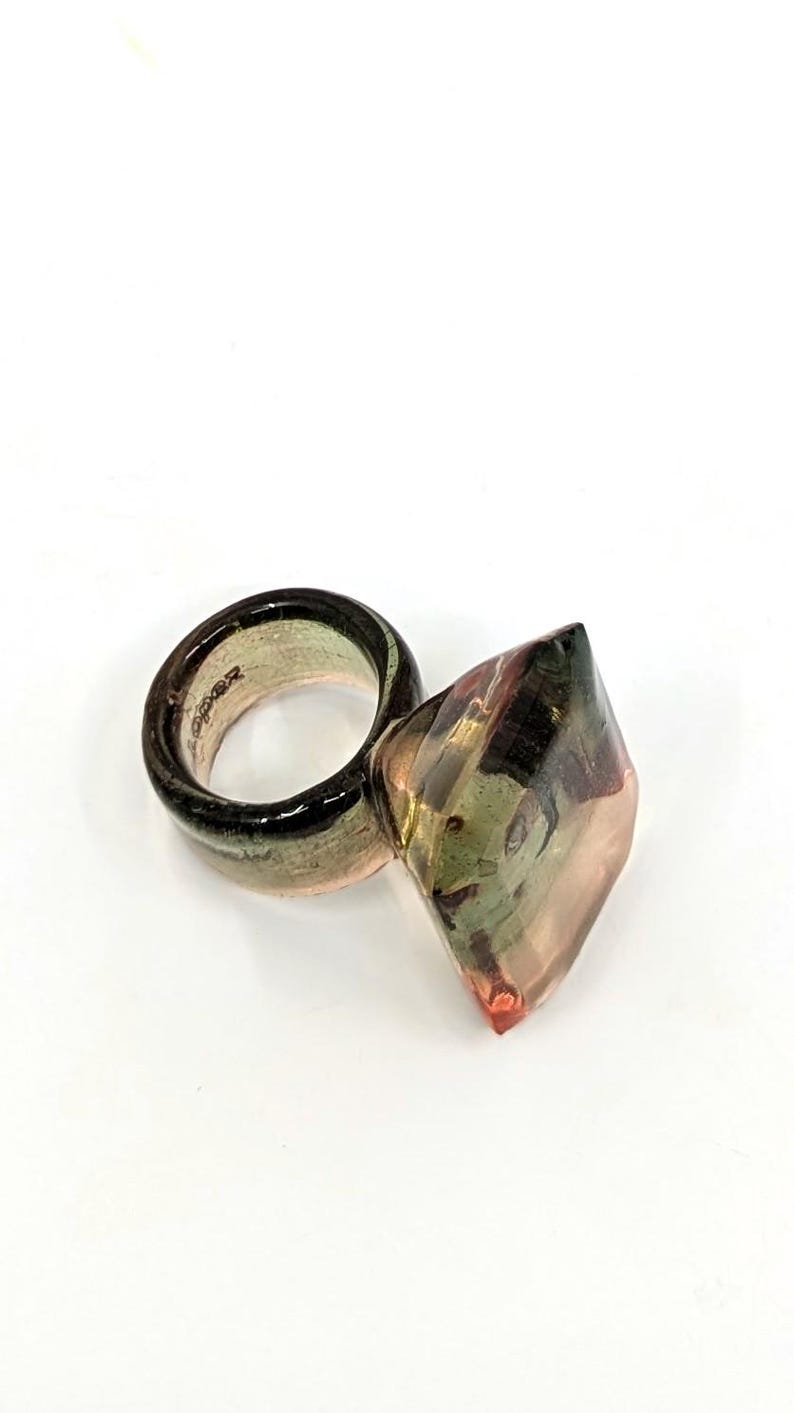 May include: A ring with a large, faceted, clear, green, and pink gemstone. The ring band is a thick, clear, green, and pink band.