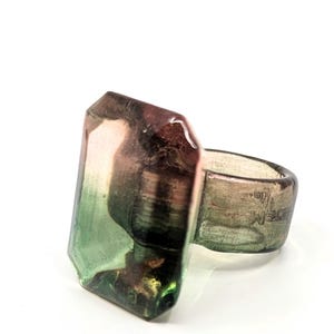 May include: A ring with a large, faceted, green, pink, and black gemstone. The ring band is made of a light brown material.