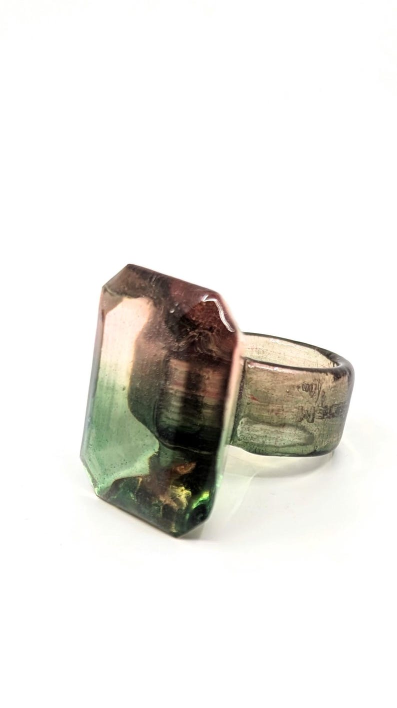 May include: A ring with a large, faceted, green, pink, and black gemstone. The ring band is made of a light brown material.