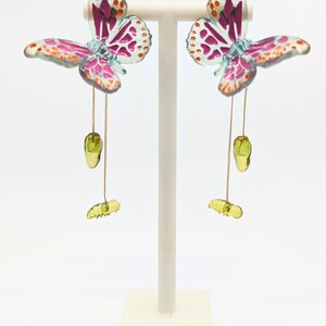 May include: A pair of butterfly earrings with pink, orange, and green accents. The earrings are hanging from a gold chain and feature a green butterfly charm.