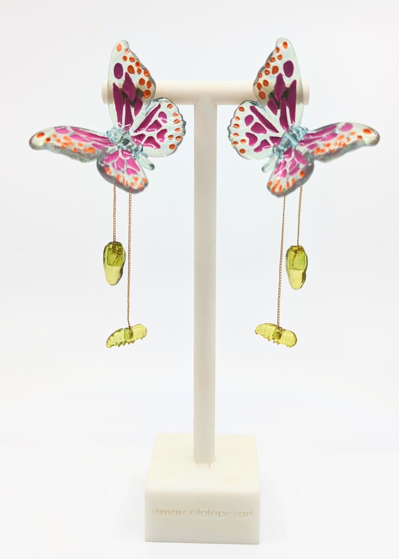 May include: A pair of butterfly earrings with pink, orange, and green accents. The earrings are hanging from a gold chain and feature a green butterfly charm.