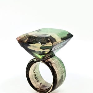 May include: A green, black, and white camouflage patterned ring with a square-shaped stone. The ring is made of wood and has a silver band.