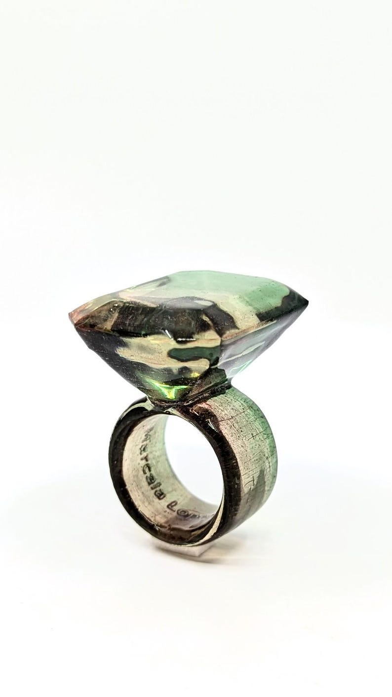 May include: A green, black, and white camouflage patterned ring with a square-shaped stone. The ring is made of wood and has a silver band.