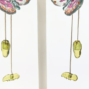 May include: A pair of dangle earrings featuring a butterfly design with pink, white, and blue accents. The earrings have a green glass charm hanging from a gold chain.