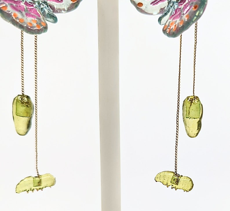 May include: A pair of dangle earrings featuring a butterfly design with pink, white, and blue accents. The earrings have a green glass charm hanging from a gold chain.