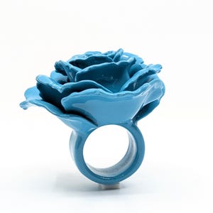 Puede incluir: Un anillo de color azul brillante con un diseño de una gran rosa esculpida en la parte superior. La banda es circular y lisa, y toda la pieza tiene un acabado uniforme de esmalte azul vibrante que refleja la luz en su superficie.