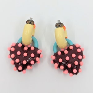 Puede incluir: Un par de pendientes con un aro amarillo y un dije en forma de corazón. El dije en forma de corazón está cubierto de puntos rosas y tiene un acento azul.