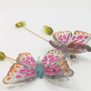 May include: A pair of dangle earrings featuring a pink and orange butterfly design with clear wings and a green bead accent. The earrings are made of resin and have a gold-tone finish.
