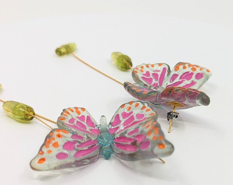 May include: A pair of dangle earrings featuring a pink and orange butterfly design with clear wings and a green bead accent. The earrings are made of resin and have a gold-tone finish.