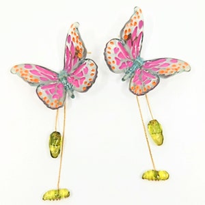 May include: A pair of dangle earrings featuring pink and orange butterfly charms with green chrysalis accents hanging from gold chains.