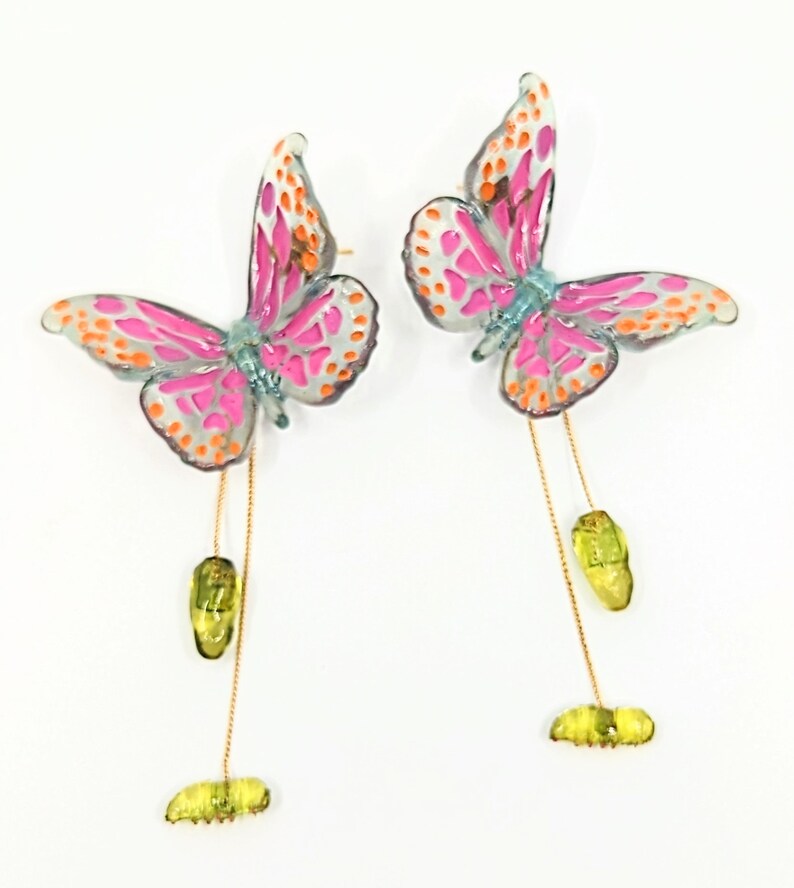 May include: A pair of dangle earrings featuring pink and orange butterfly charms with green chrysalis accents hanging from gold chains.