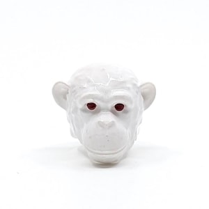 Puede incluir: Una cabeza de mono de cerámica blanca con ojos rojos. Este objeto decorativo es probablemente una figura o parte de una escultura. Los rasgos faciales del mono están bien definidos.