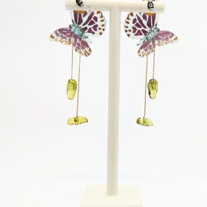 May include: A pair of dangle earrings featuring a butterfly design with purple, blue, and gold accents. The butterflies are made of clear plastic with a spotted pattern. The earrings hang from a gold chain with a green stone charm.
