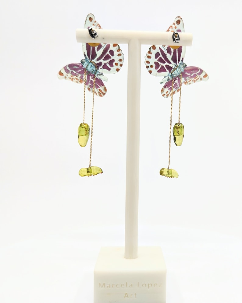May include: A pair of dangle earrings featuring a butterfly design with purple, blue, and gold accents. The butterflies are made of clear plastic with a spotted pattern. The earrings hang from a gold chain with a green stone charm.