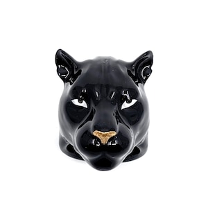 Puede incluir: Un anillo de cerámica negra brillante con forma de cabeza de pantera. La pantera presenta orejas puntiagudas, ojos blancos almendrados con pupilas negras y una nariz texturizada de color dorado. El aro está integrado en la base del diseño.