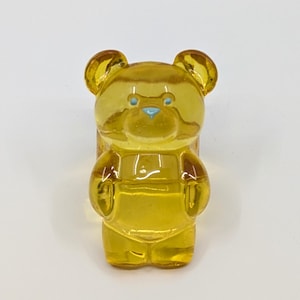 May include: A translucent yellow gummy bear ring featuring a smooth, glossy finish. The bear has small, light blue painted eyes and a light blue nose. The design is moulded into a classic bear shape with rounded ears, arms at its sides, and legs.