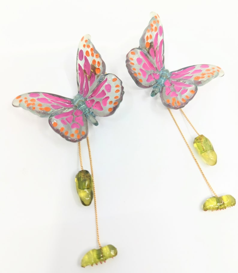 May include: A pair of butterfly earrings with pink, orange, and blue accents. Each butterfly has a gold chain with a green chrysalis and a green caterpillar dangling from it.
