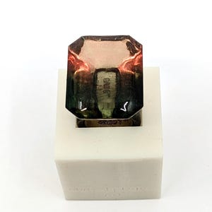 May include: A clear, multi-colored, square-shaped ring with a green, brown, and pink gradient. The ring is sitting on a white square base with the words "Made in Italy" printed on the base.