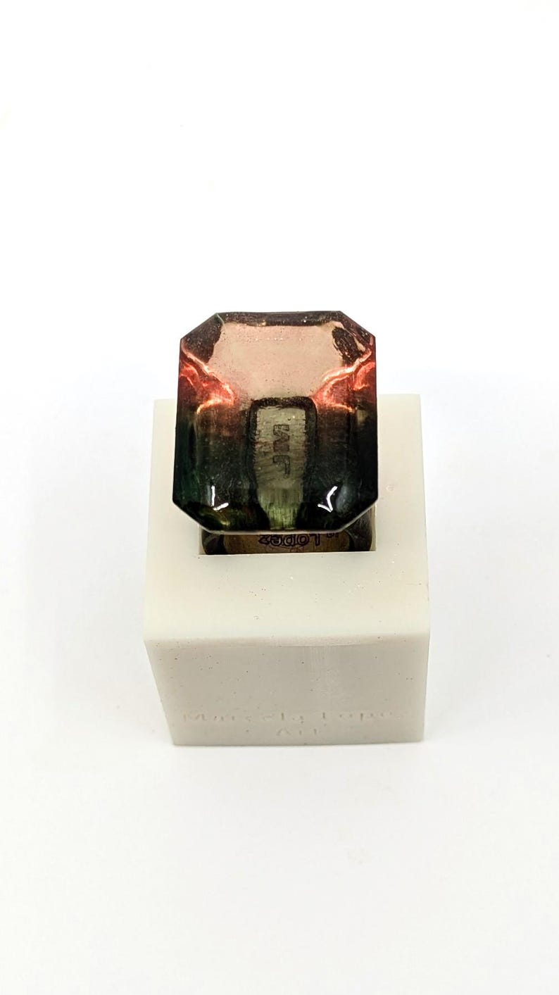 May include: A clear, multi-colored, square-shaped ring with a green, brown, and pink gradient. The ring is sitting on a white square base with the words "Made in Italy" printed on the base.