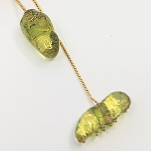 May include: A gold chain necklace with a green glass caterpillar charm and a green glass chrysalis charm.