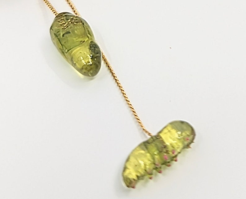 May include: A gold chain necklace with a green glass caterpillar charm and a green glass chrysalis charm.