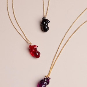 Puede incluir: Tres collares de oro con colgantes de corazón anatómico. Los corazones están hechos de vidrio transparente y están disponibles en negro, rojo y morado.
