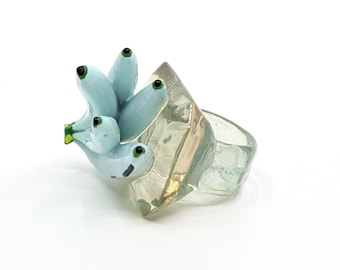 May include: A clear plastic ring with a blue and green glass banana charm. The ring is shaped like a square with rounded edges.