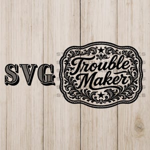 May include: Black SVG design featuring the text "SVG" and "Trouble Maker." The words are in a decorative font. "Trouble Maker" is inside a detailed, ornate frame with stars and flourishes. The background is a light wood grain.