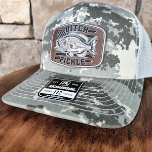 May include: A camouflage-patterned trucker hat with a brown leather patch featuring a fish graphic and the words "Ditch Pickle." The hat has a white mesh back and a curved brim. The Richardson brand label is visible.