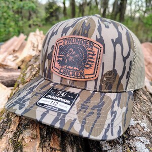 May include: A camouflage-patterned Richardson 112 trucker hat with a brown leather patch featuring a turkey graphic and the words "THUNDER CHICKEN". The hat has a mesh back and an adjustable strap.