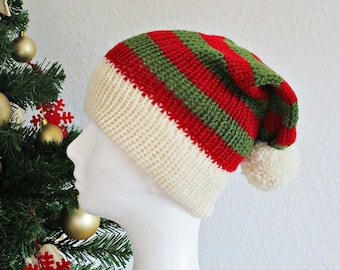 May include: A knitted beanie featuring horizontal red and green stripes with a wide cream-colored ribbed cuff and a fluffy white pom-pom at the tip. The hat is displayed on a white mannequin head, showcasing the textured yarn construction.
