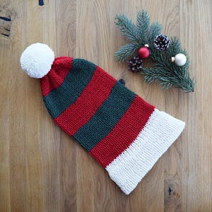 Könnte beinhalten: Eine gestrickte Weihnachtsstrumpfmütze mit roten, grünen und weißen Streifen, obenauf ein weißer Bommel. Die Mütze liegt auf einer Holzoberfläche, mit einem Zweig, Tannenzapfen und Ornamenten.