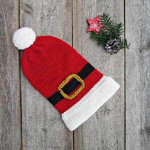 Könnte beinhalten: Eine rote gestrickte Weihnachtsmütze mit weißem Bommel und Krempe. Die Mütze hat ein schwarzes Band mit einer goldfarbenen Schnalle. Festliche Weihnachtsdekoration.