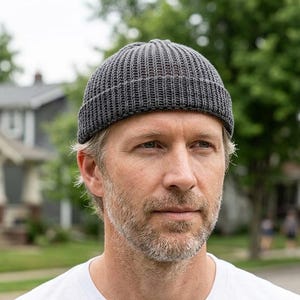 Könnte beinhalten: Eine graue, gerippte Strickmütze. Die Mütze wird von einer Person in einem weißen T-Shirt getragen. Der Hintergrund zeigt eine Wohnstraße mit Häusern und Bäumen.